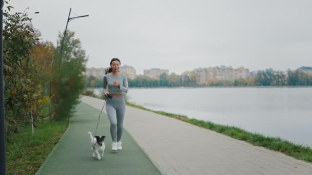 Női futár és a kutyája a városi tó közelében reggelente. Szabadtéri séta. Atlétikai fiatal lány és kutyája fut a városban - Felvétel, videó