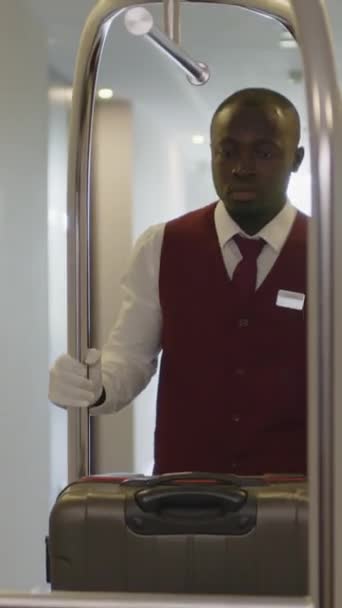Colpo verticale del giovane fattorino afroamericano in uniforme che attraversa il corridoio dell'hotel e porta bagagli sul carrello - Filmati, video