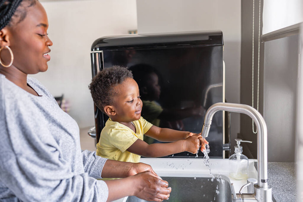 Cute little smiling African-American boy washing hands in a kitchen sink at home - Photo, Image