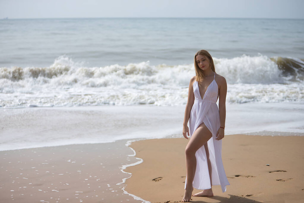 Jonge, mooie, blonde vrouw in witte jurk, poserend op een eenzaam strand, ontspannen en kalm. Concept vrede, rust, eenzaamheid, tederheid, maagdelijkheid. - Foto, afbeelding