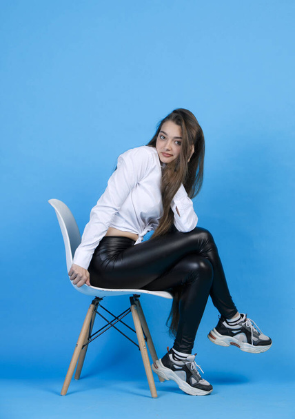Pretty caucasian woman in white blouse, resting head on hand, while sitting on chair cross legged. Side view of cute girl smiling, while posing at camera, isolated on blue. Concept of female posing. - Photo, Image