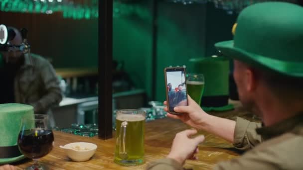 Через плечо неопознанные мужчина и женщина в зеленых шляпах звонят ирландским друзьям и поздравляют с Днем Святого Патрика по видеосвязи во время празднования у стойки в баре с зеленым пивом в пабе - Кадры, видео