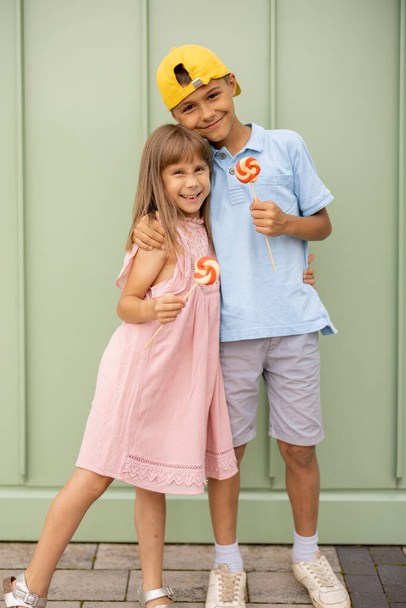 Portrait of a happy boy with his little sister stand with sweet candies on green wall background outdoors, spending summertime together. Little boy with a girl visiting amusement park and having fun - Photo, Image