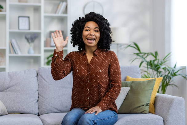 Retrato de una joven afroamericana sonriente sentada en el sofá de su casa frente a la cámara, saludando y saludando. - Foto, imagen