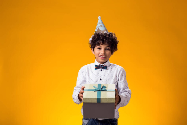 Portrait of child boy with birthday cap on head holding giftbox over yellow background. Birthday and celebration concept - Photo, Image