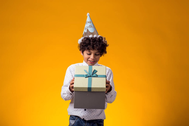 Портрет хлопчика з шапочкою для дня народження на голові, що тримає подарункову коробку на жовтому тлі. Концепція дня народження та святкування - Фото, зображення