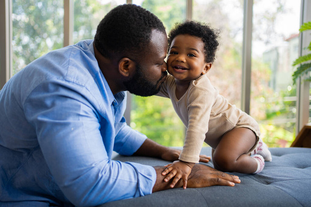 Portrait Of Happy African American Dad With Cute Little Baby Girl auf der Couch zu Hause im Wohnzimmer, fürsorglicher Vater lächelt und amüsiert sein Mädchen, während es auf der Couch sitzt, glückliche Familie - Foto, Bild