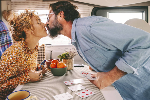 Couple enjoy leisure time playing cards and kissing with love inside a camper van. Travel and holiday vacation concept. Van life for travel people. Young adult man and woman in relationship indoor - Photo, Image
