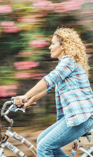 Щаслива доросла жінка катається на велосипеді на відкритому повітрі з квітами на задньому плані. Навколишнє середовище та зелений транспорт з велосипедом. Веселі сучасні жінки насолоджуються активним дозвіллям, катаючись з посмішкою
 - Фото, зображення