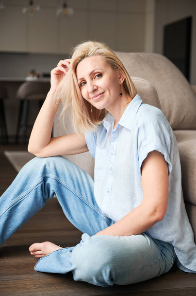 Портрет красивой женщины с светлыми волосами, расслабляющей рядом с диваном. Женщина сидит на деревянном полу. - Фото, изображение