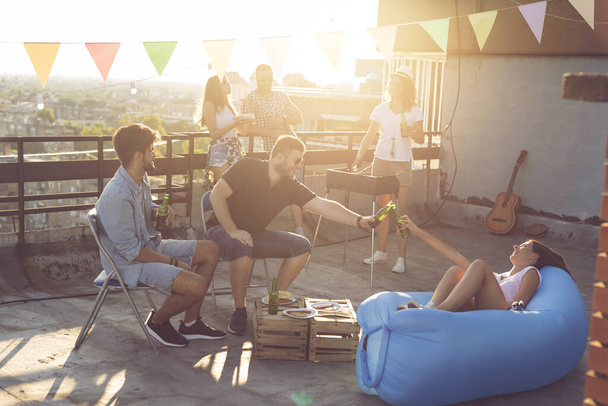 Groep jonge vrienden die zich vermaken op een feestje op het dak, barbecueën, bier drinken en genieten van warme zomerdagen. Focus op de mensen op de voorgrond - Foto, afbeelding