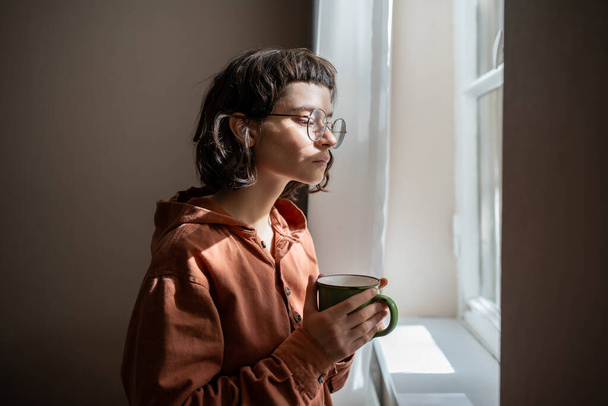 Девушка-ботаник в очках смотрит в окно и пьет чай дома. Серьёзная молодая женщина отдыхает, созерцая улицу, наблюдая за людьми. Уикенд, интроверт, отдых в концепции одиночества. - Фото, изображение