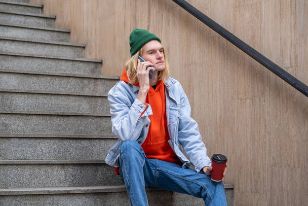 Elinde kahve fincanı olan sakin hipster adam merdivenlerde oturup şehir caddesinde akıllı telefondan konuşuyor. Şehir merkezinde uzun bir yürüyüşten sonra dinlenen bir adam, dışarıda merdivenlerde oturuyor..  - Fotoğraf, Görsel