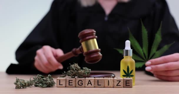 Word Legalize made of wooden cubes in front of judge with gavel. Woman holds fresh marijuana leaf near bottle with psychoactive drug substance - Footage, Video