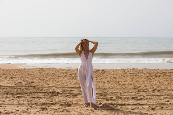 Молодая красивая блондинка в белом платье гуляет по песку на берегу пляжа в солнечный день. Женщина делает различные выражения тела. На заднем плане голубое море. - Фото, изображение