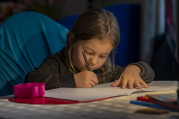 Portret van een blond meisje dat 's avonds studeert onder het licht van een tafellamp. - Foto, afbeelding
