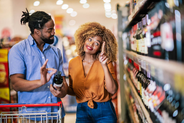 Afrikkalainen amerikkalainen pari, jolla on kärry ostamassa ostoksia ostoskeskuksesta. Viinin ostaminen. - Valokuva, kuva