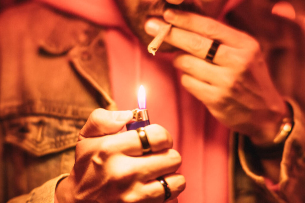 Yksityiskohta miehen käsistä sytyttämässä marihuanasavuketta punaisen valon alla. - Valokuva, kuva