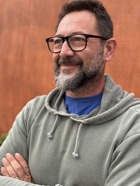 Retrato del hombre adulto sonriendo y mirando al frente con gafas y suéter verde contra un fondo de pared marrón en la actividad de ocio al aire libre solo - Foto, imagen