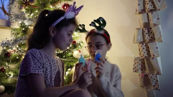 Twee mooie blanke meisjes gelukkig eten snoep en spelen met speelgoed van een muur adventskalender cadeau, zitten in de avond tegen de achtergrond van een elegante kerstboom, close-up side view. - Video