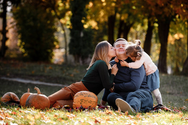 Ημέρα των Ευχαριστιών. Χαρούμενη μαμά, ο μπαμπάς και η μικρή κόρη διασκεδάζουν κάθονται στο γρασίδι με κολοκύθες στο πάρκο φθινόπωρο, απολαμβάνοντας περνούν το χρόνο τους μαζί το Σαββατοκύριακο. Γονική φροντίδα και ευτυχισμένη ανέμελη παιδική ηλικία. - Φωτογραφία, εικόνα