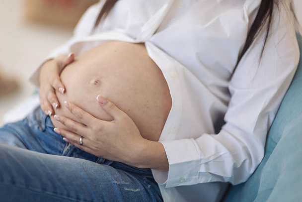 Těhotná žena v bílé košili uvnitř, detailní záběr. těhotná žena drží ruce na svém oteklém břiše. koncepce očekávání těhotenství. - Fotografie, Obrázek