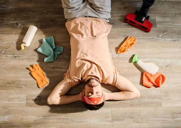Draufsicht auf einen jungen müden Mann in Freizeitkleidung und Stirnband, der mit Staubsauger, Staubwedel, Reinigungsmitteln und Handschuhen auf sauberem Boden liegt - Foto, Bild