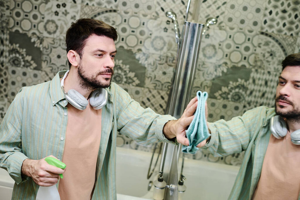Hombre en ropa casual sosteniendo botella de detergente con pulverizador y espejo limpiador en el baño con plumero húmedo durante las tareas domésticas diarias - Foto, Imagen