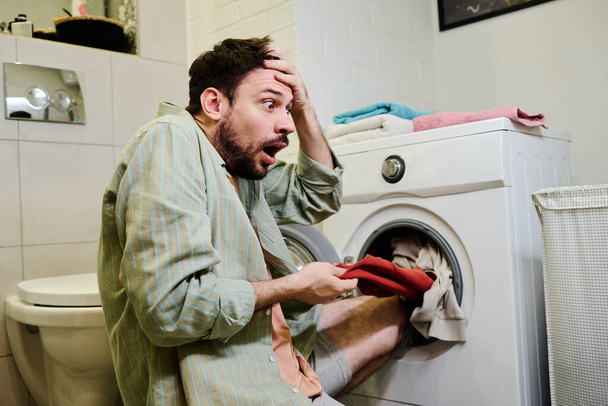 Geschokt jongeman trekken rode stuk van kleding uit wasmachine en kijken naar het met doodsbang uitdrukking terwijl het aanraken van zijn hoofd - Foto, afbeelding
