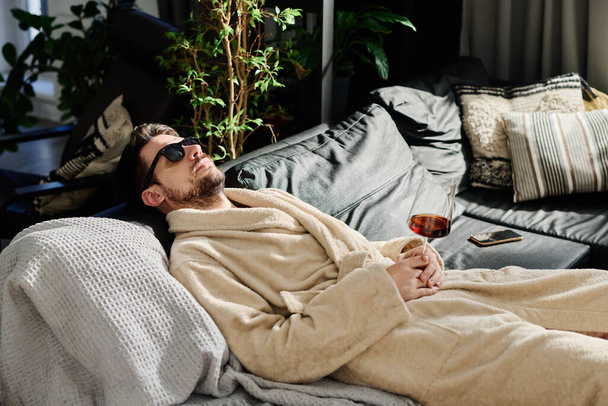Ξεκούραστος τύπος με μπουρνούζι και γυαλιά ηλίου κρατώντας ένα ποτήρι κόκκινο κρασί στο χέρι ενώ χαλαρώνει στον καναπέ με μαξιλάρια και ονειροπολεί - Φωτογραφία, εικόνα