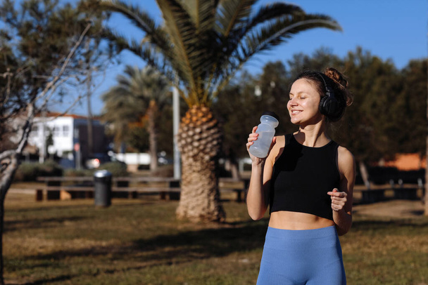 Active young fit woman in wireless headphones drinks water from bottle after exercising outdoors at palm trees park. Happy girl in sportswear quenches thirst and cools down after running or workout. - Photo, Image