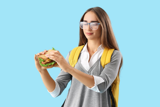 Νεαρή φοιτήτρια τρώει νόστιμο σάντουιτς σε μπλε φόντο - Φωτογραφία, εικόνα