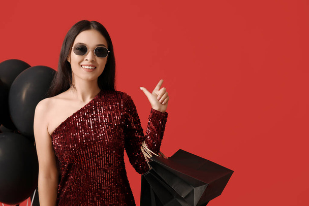 Hermosa mujer asiática joven con bolsas de compras y globos apuntando a algo en el fondo rojo. Viernes Negro venta - Foto, Imagen
