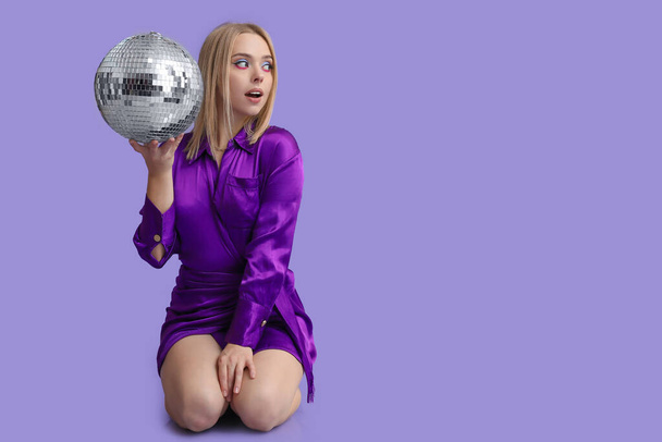 Beautiful shocked young woman with disco ball sitting on purple background - Photo, Image