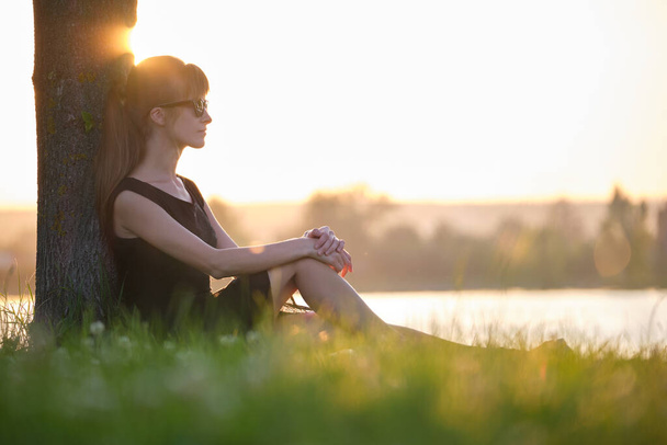 Nuori nainen rento asu istuu vihreä ruoho nurmikko rentouttava järven puolella lämpimänä iltana. Kesälomat ja matkakonsepti. - Valokuva, kuva