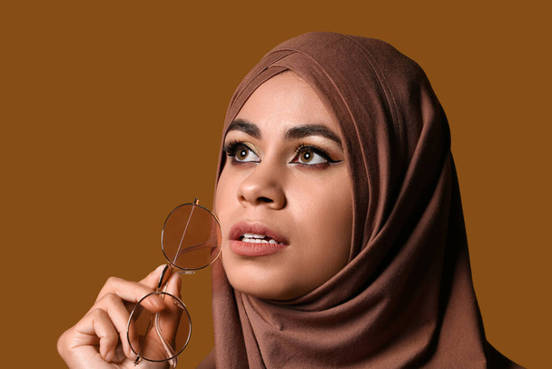 Молодая афроамериканка-мусульманка с красивым макияжем и стильными солнечными очками на коричневом фоне - Фото, изображение