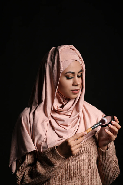 Joven mujer musulmana afroamericana con hermoso maquillaje sosteniendo polvo facial y cepillo sobre fondo negro - Foto, imagen