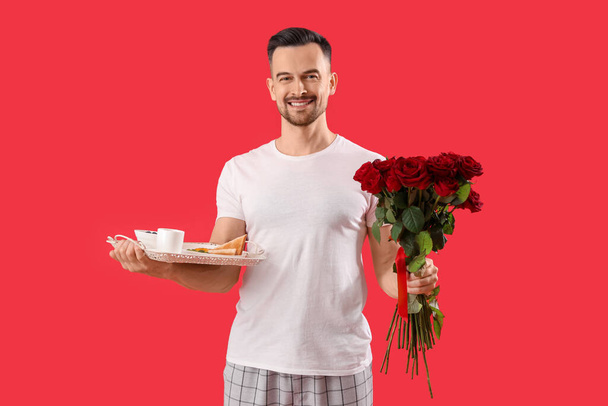 Junger Mann mit leckerem Frühstück und Rosen auf rotem Hintergrund. Valentinstag-Feier - Foto, Bild
