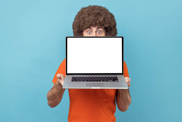Anonymous man with Afro hairstyle in orange T-shirt holding laptop in hands, hiding half of his face, copy space for promotion, looking at camera. Indoor studio shot isolated on blue background. - Фото, изображение