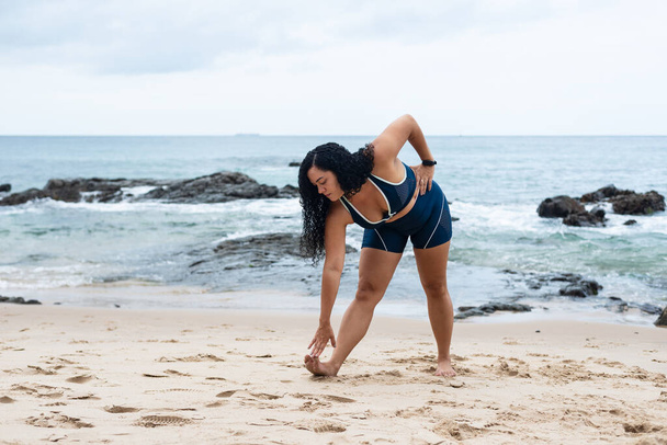 Piękna, kręcone włosy, młoda kobieta w stroju siłowni robi rozciąga się na piasku plaży. Zdrowy styl życia. - Zdjęcie, obraz