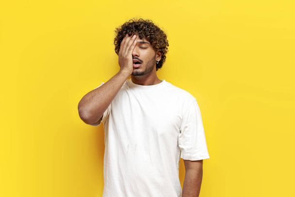 giovane uomo indiano riccio mostrando facepalm e fallimento su sfondo giallo isolato, ragazzo triste ha perso e coprendo il viso con la mano da fare un errore - Foto, immagini