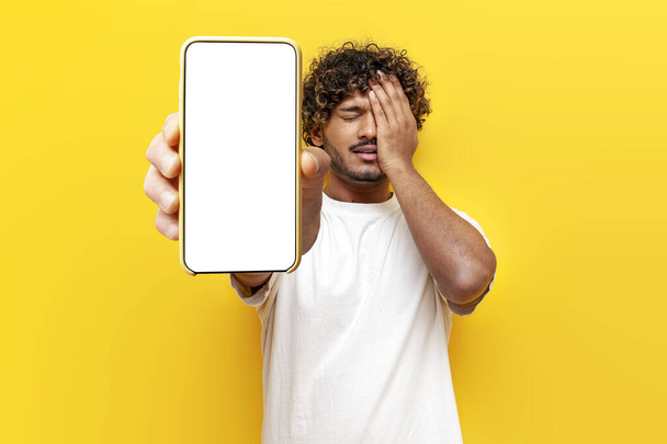 malheureux homme indien montrant écran vide de smartphone et se sentir triste par erreur et l'échec sur fond jaune isolé, gars perd en ligne et montre facepalm - Photo, image