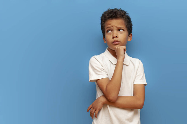 african american sad teenager boy in white polo thinks and plans on blue isolated background, unhappy pensive child dreams and imagines - Photo, Image