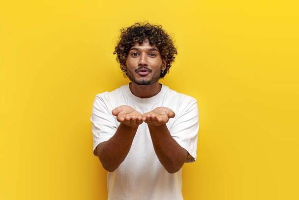 jonge krullende indiaanse man houdt lege handen voor zich en stuurt een luchtkus op gele geïsoleerde achtergrond, man toont romantisch gebaar en flirten en blazen op handen - Foto, afbeelding