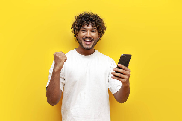 młody kręcone indyjski facet za pomocą smartfona i świętowanie zwycięstwo na żółtym odizolowanym tle, facet radujący się szczęście online i wygrywając na smartfonie - Zdjęcie, obraz