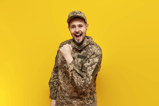 young Ukrainian male soldier in pixel camouflage uniform celebrates victory on a yellow isolated background, guy military cadet of the Ukrainian army wins and rejoices at luck and success - Photo, Image