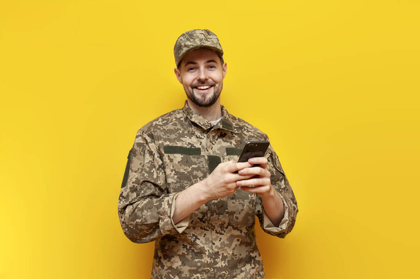un jeune soldat ukrainien en uniforme de camouflage pixel utilise un smartphone sur un fond jaune isolé, un cadet militaire de l'armée ukrainienne communique en ligne - Photo, image