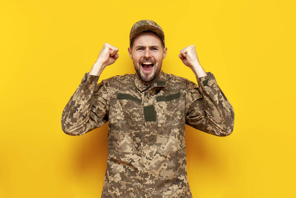 молодий український військовослужбовець у піксельній камуфляжній формі відзначає перемогу на жовтому ізольованому тлі, хлопець-військовий кадет української армії перемагає і радіє удачі та успіху - Фото, зображення