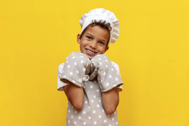 garçon afro-américain boulanger en uniforme et chef chapeau sur fond jaune isolé, adolescent enfant en gants thermiques de cuisson regardant la caméra - Photo, image