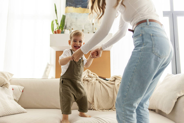 giovane madre e il suo piccolo figlio ballare e saltare sul divano a casa, 2-year-old boy urla e gioisce con il suo genitore, la donna si diverte con il suo bambino - Foto, immagini